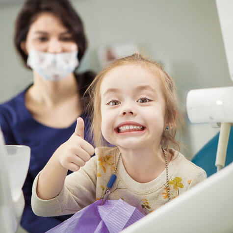 Young girl brushing teeth with dental hygienist in mask supervising at pediatric dental office.