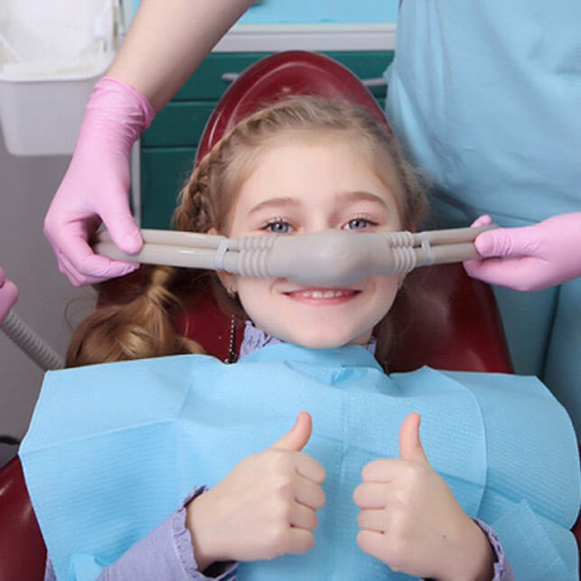 Young child at dental office giving thumbs up while dentist measures mouth with dental tool.