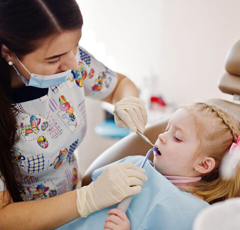 Pediatric dentist in mask examining young child's teeth in dental office